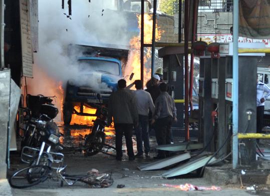 Syrian citizens stand near a burning truck that was destroyed by two cars bombs. (AP/SANA)