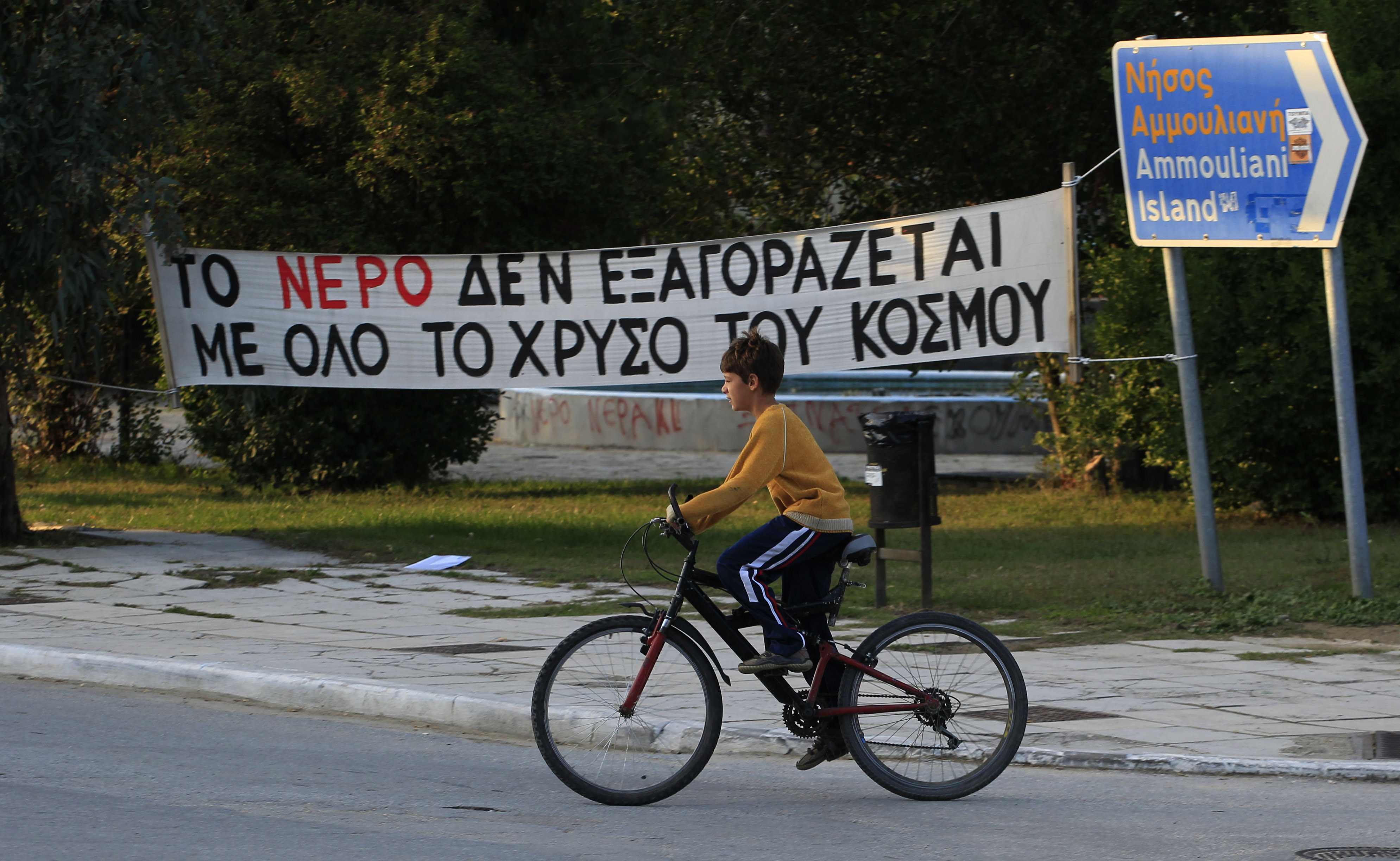 A boy cycles past a banner which reads in Greek "All the gold in the world can't buy water" at the village of Ierissos in the Halkidiki peninsula in northern Greece. (AP/Nikolas Giakoumidis)