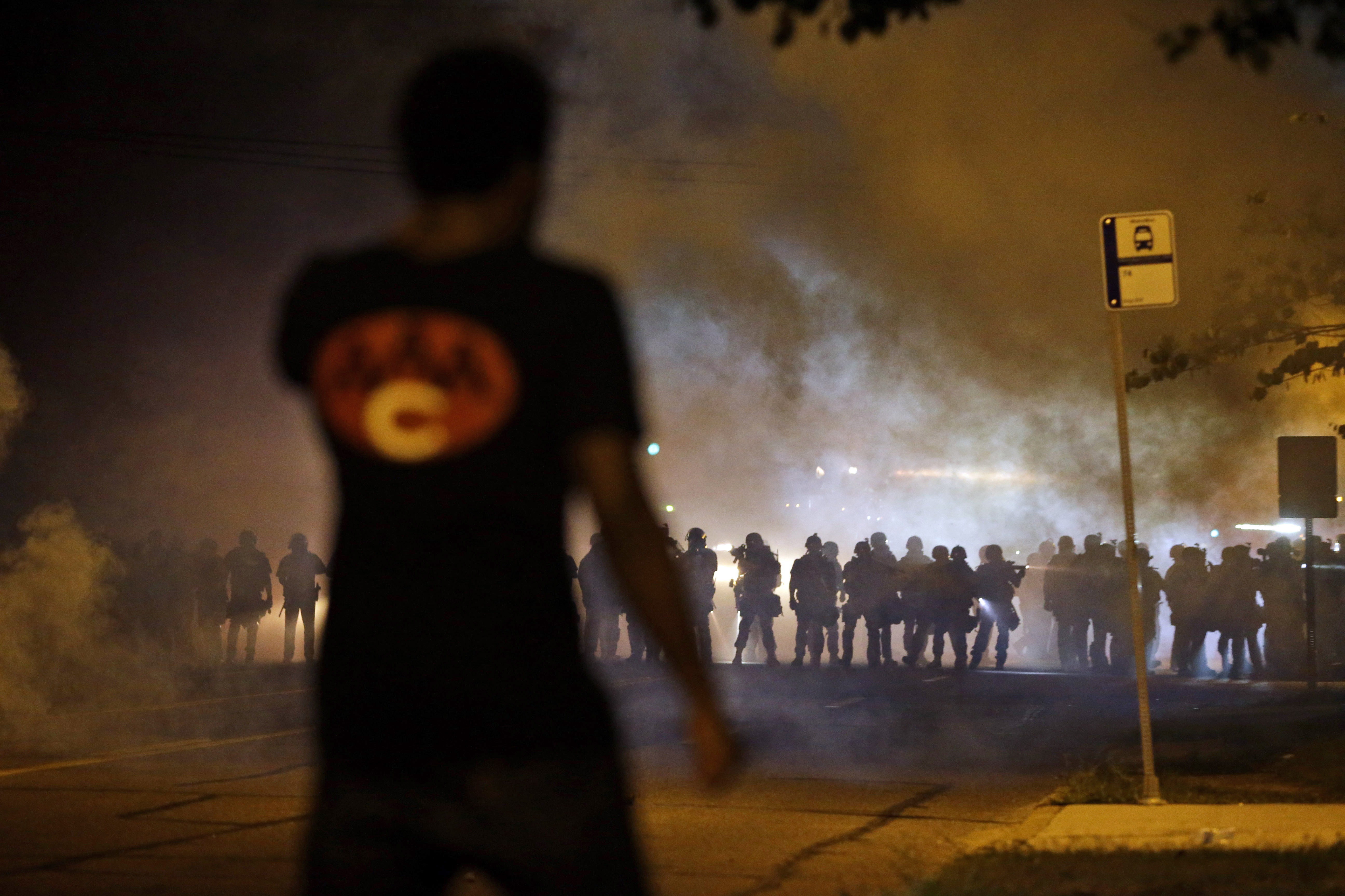 FILE - In this Aug. 13, 2014 file photo, a man watches as police walk through a cloud of smoke during a clash with protesters in Ferguson, Mo. The response to Browns death turned violent because of a convergence of factors, observers said. (AP/Jeff Roberson)