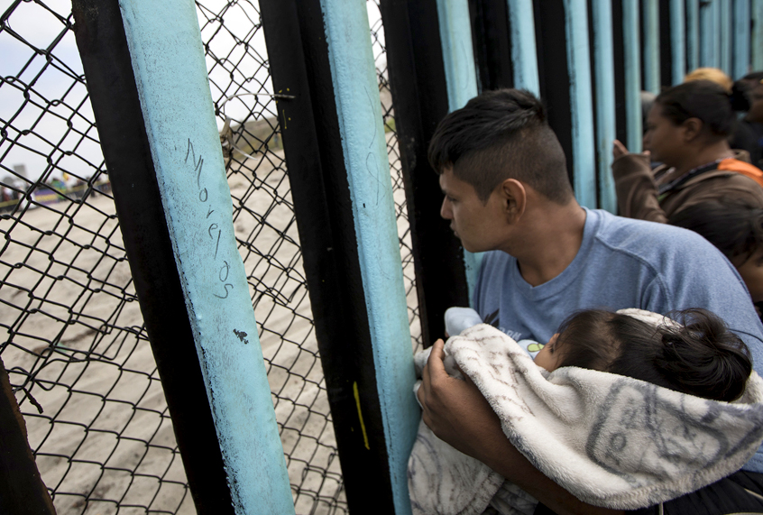 A member of the Central American migrant caravan, holding a child, looks through the U.S./Mexico border wall. (AP/Hans-Maximo Musielik)