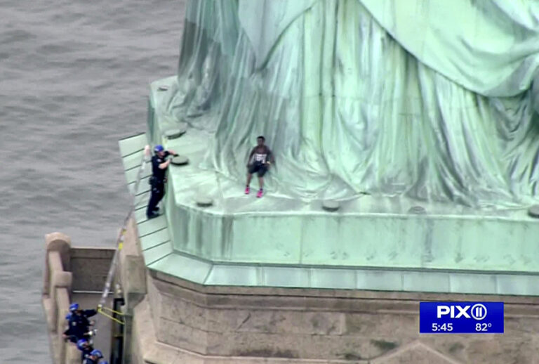 In this image made from video by PIX11, a person, center, leans against the robes of the Statue of Liberty on Liberty Island, as one of the police officers climbed up on a ladder to stand on a ledge nearby talking the climber into descending in New York, Wednesday, July 4, 2018. (PIX11 via AP)