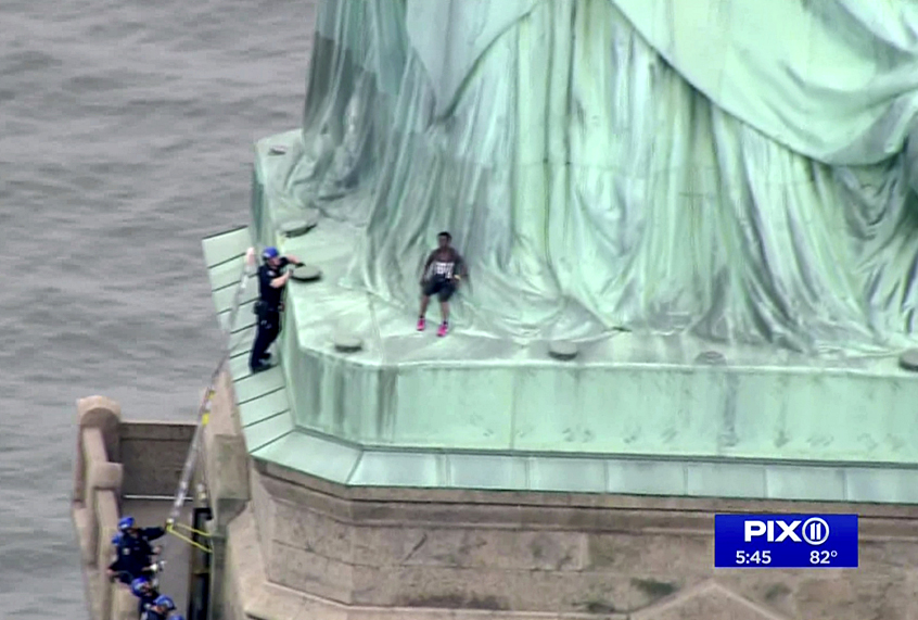 In this image made from video by PIX11, a person, center, leans against the robes of the Statue of Liberty on Liberty Island, as one of the police officers climbed up on a ladder to stand on a ledge nearby talking the climber into descending in New York, Wednesday, July 4, 2018. (PIX11 via AP)