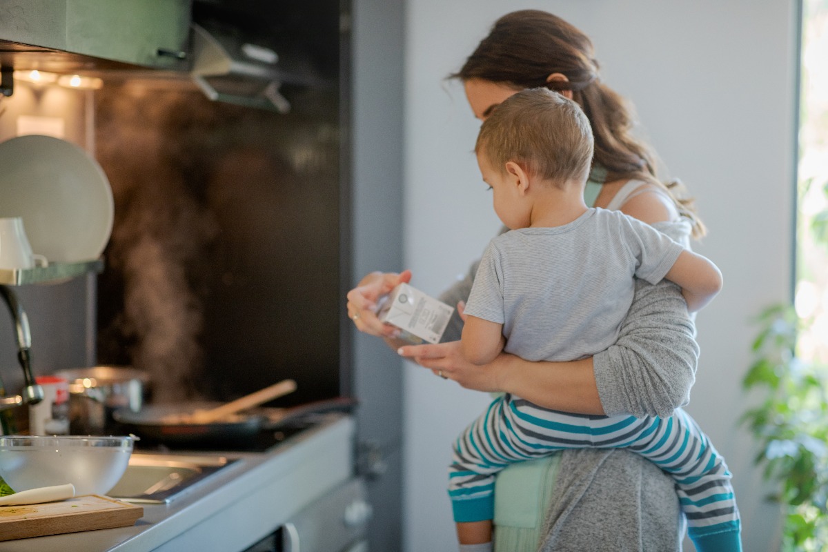 Mother with toddler on hip holding a carton of cooking cream and checking label. (Getty Images)