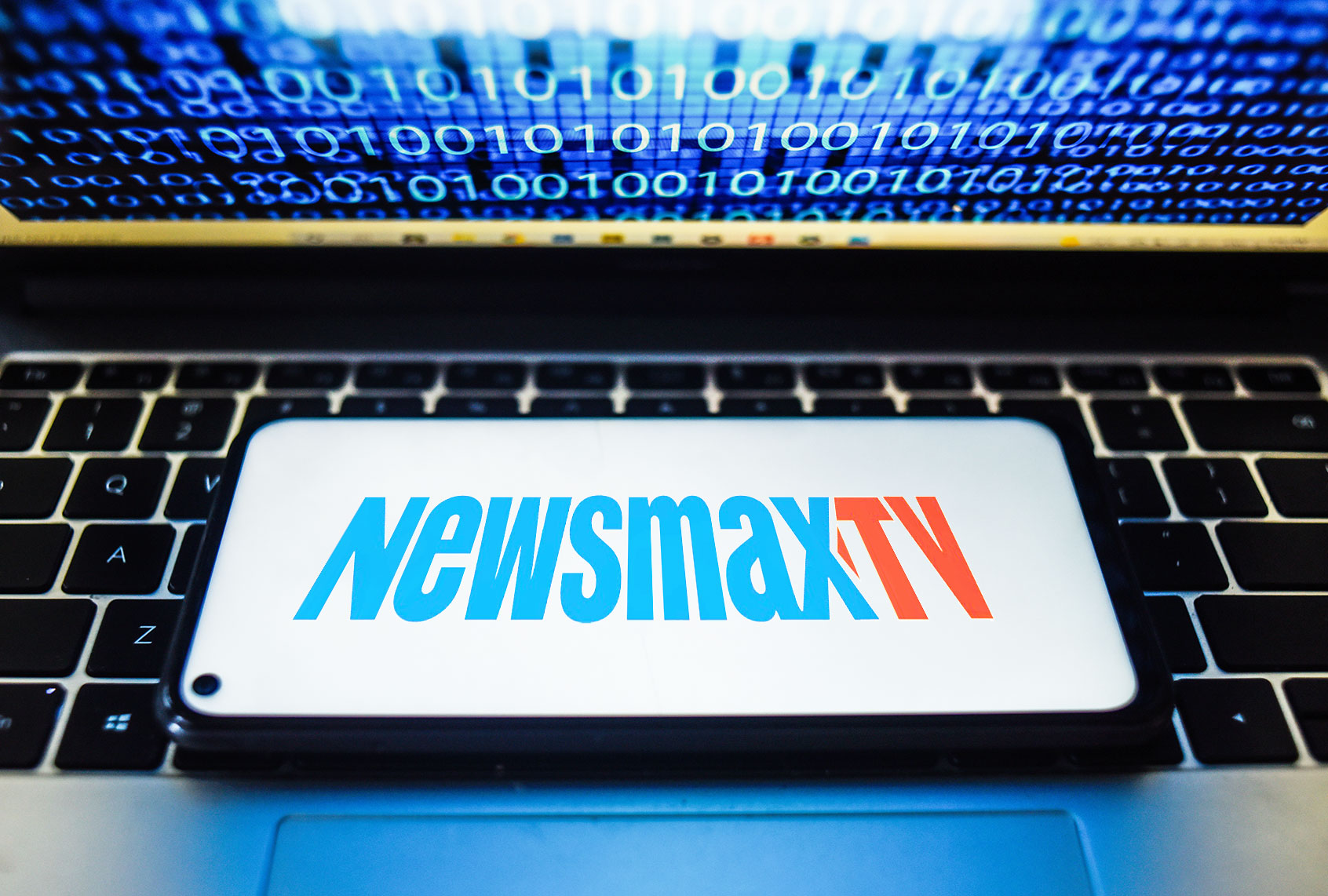 Newsmax TV logo seen displayed on a smartphone with binary code on a laptop screen. (Photo Illustration by Omar Marques/SOPA Images/LightRocket via Getty Images (Photo illustration by Salon/Omar Marques/SOPA Images/LightRocket/Getty Images/WikiCommons)