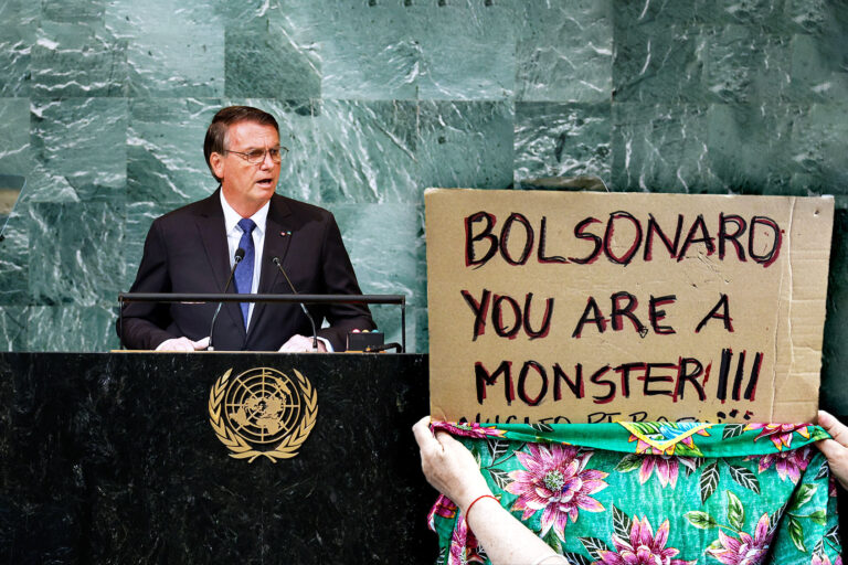Brazilian President Jair Bolsonaro speaks during the 77th session of the United Nations General Assembly (UNGA) at U.N. headquarters | Protest sign (Photo illustration by Salon/Getty Images)