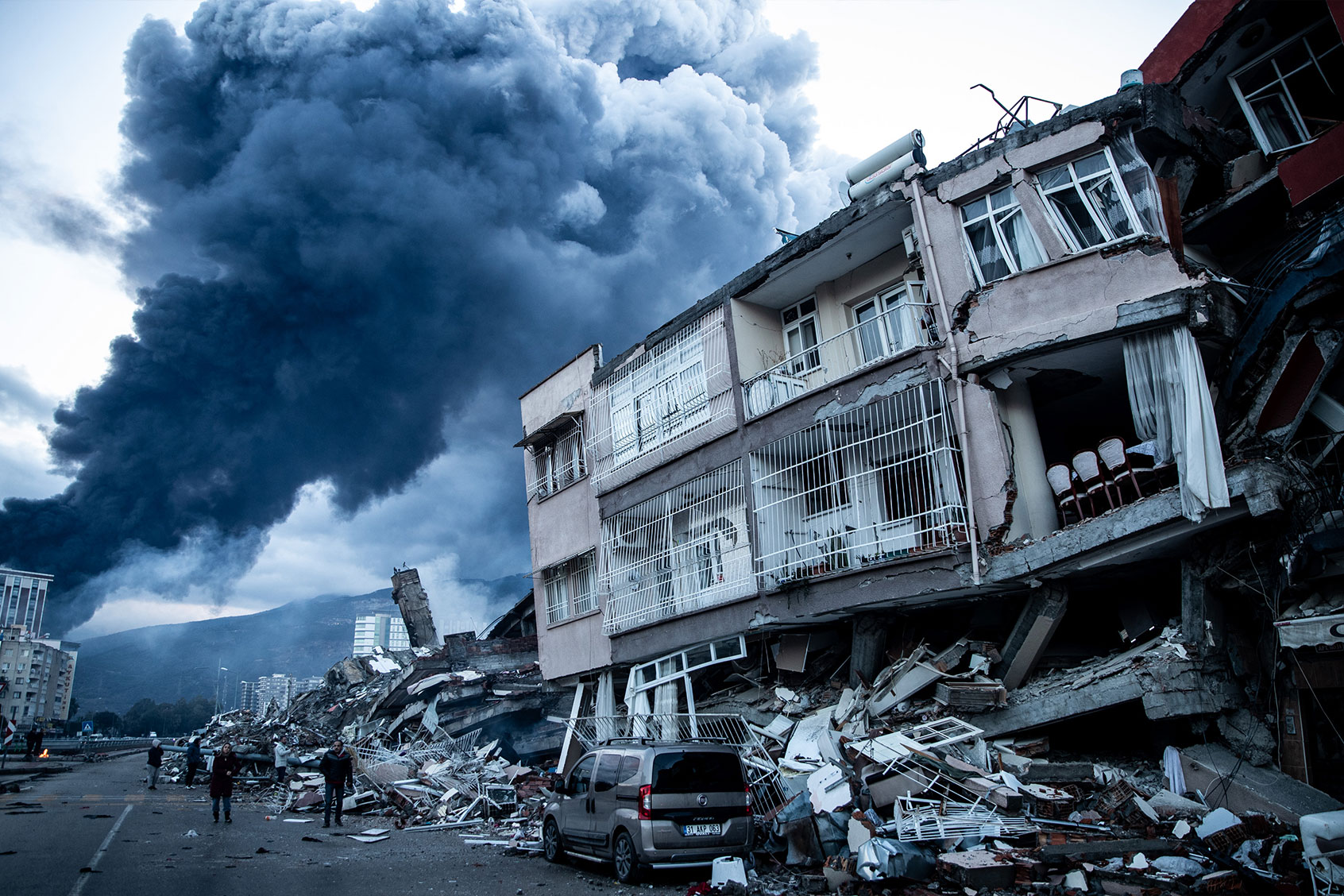 Smoke billows from Iskenderun Port fire as people walk past collapsed buildings on February 07, 2023 in Iskenderun, Turkey. (Burak Kara/Getty Images)