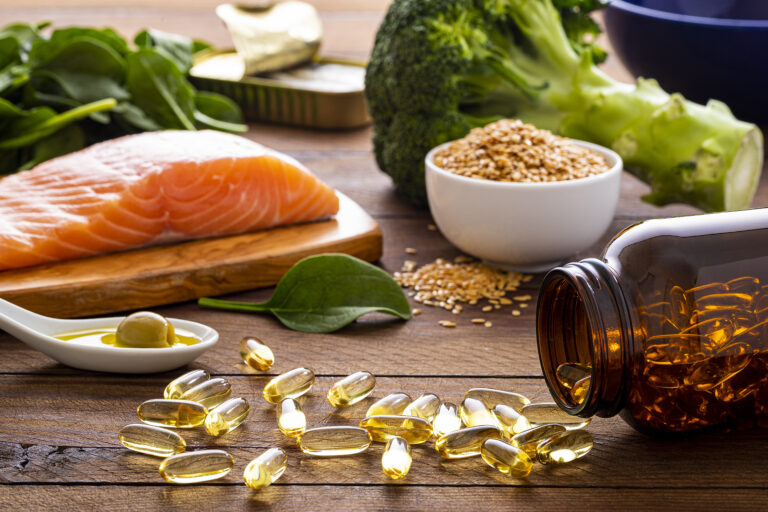 Front view of fish oil capsules spilling out from a bottle surrounded by an assortment of food rich in omega-3 such as salmon, flax seeds, broccoli, sardines, spinach, olives and olive oil. (carlosgaw / Getty Images)