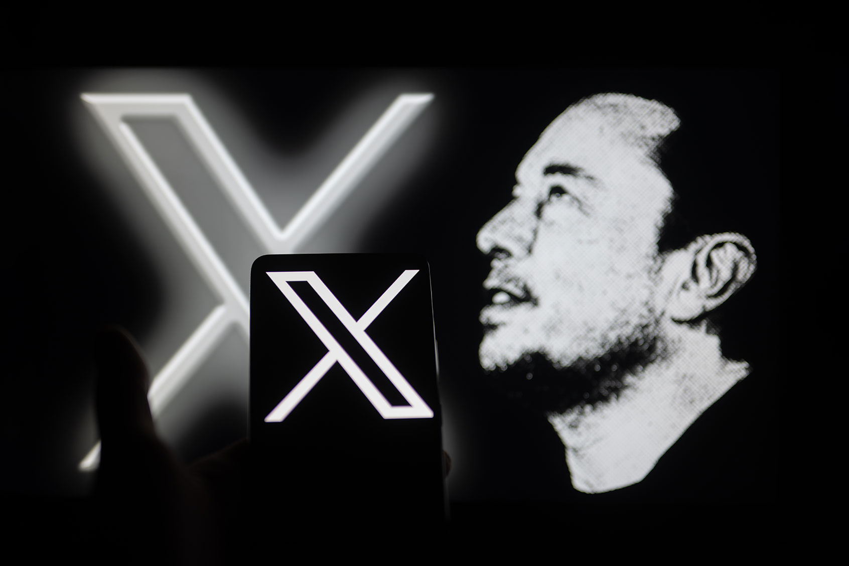 The new Twitter logo, rebranded as X, is being displayed on a smartphone with Elon Musk visible in the background, in this photo illustration in Brussels, Belgium, on November 27, 2023. (Jonathan Raa/NurPhoto via Getty Images)