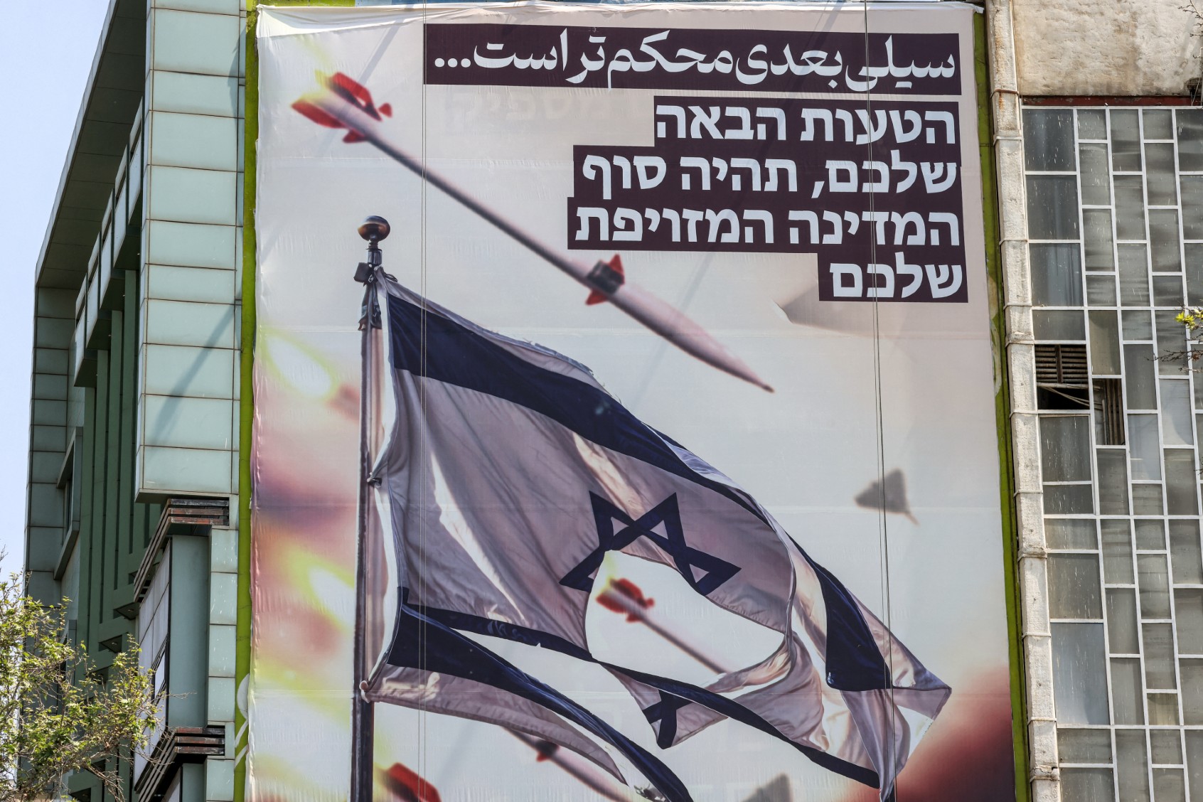 A banner depicting missiles and drones flying past a torn Israeli flag, with text in Persian reading "the next slap will be harder" and in Hebrew "your next mistake will be the end of your fake state", hangs on the facade of a building in Palestine Square in Tehran on April 14, 2024. (ATTA KENARE/AFP via Getty Images)