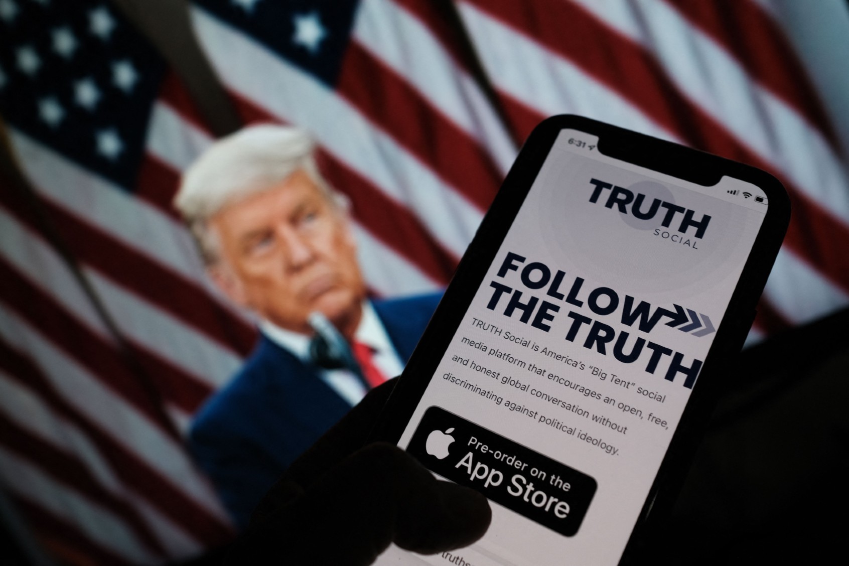 This illustration photo shows a person checking the app store on a smartphone for "Truth Social," with a photo of former President Donald Trump on a computer screen in the background, in Los Angeles, October 20, 2021. (CHRIS DELMAS/AFP via Getty Images)