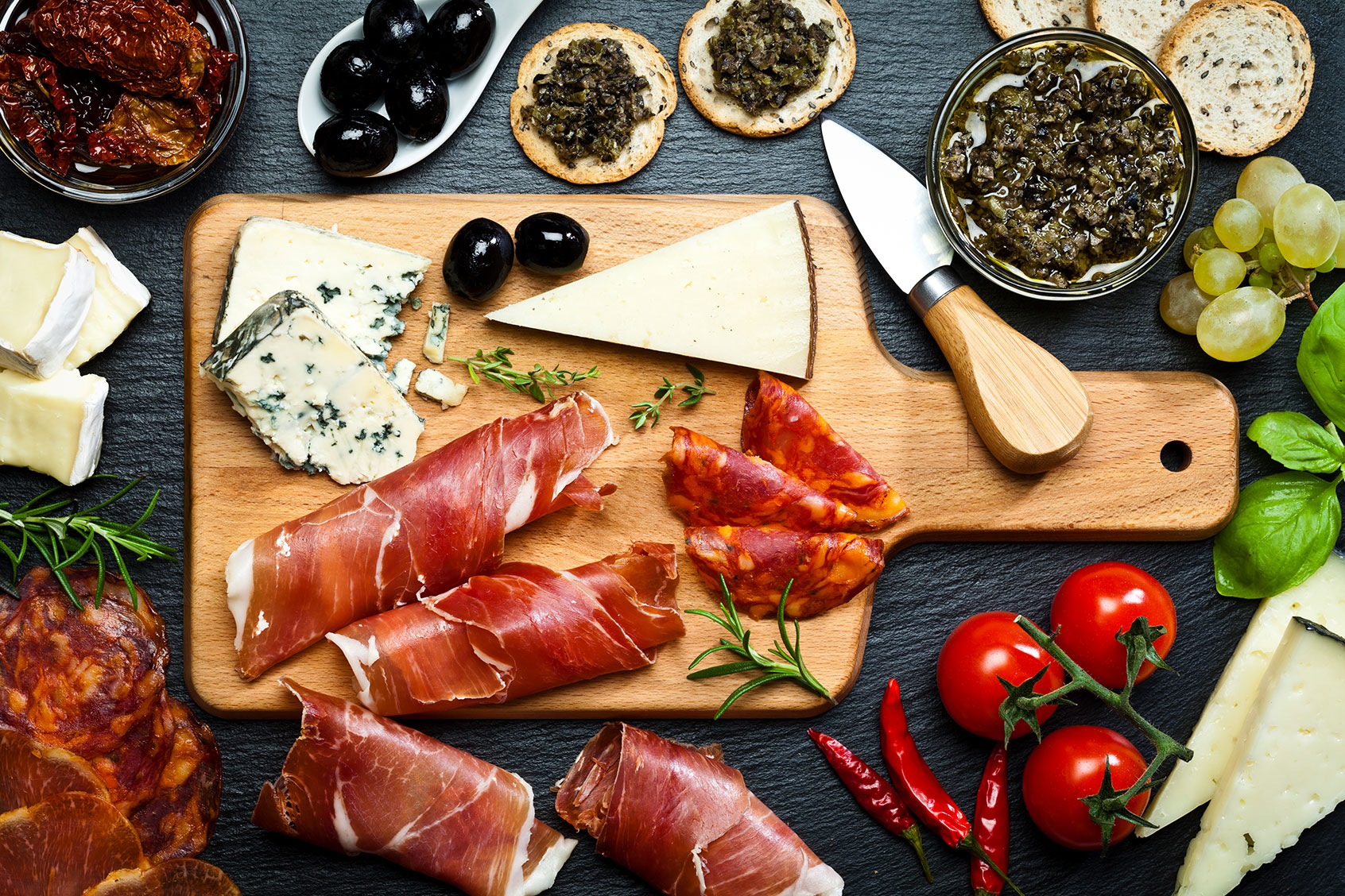 Top view of a wooden cutting board on dark slate background filled with a assortment of fine food. (Getty Images/fcafotodigital)
