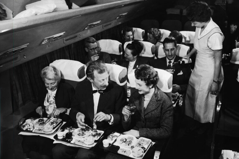 Interior view of a commercial passenger plane shows, in the foreground, a couple as they enjoy their meal next to a smiling elderly woman, while behind them, a flight attendant pours a glass of wine for a man who sits next to a couple who toast each other with full glasses, 1950s. (Frederic Lewis/Getty Images)