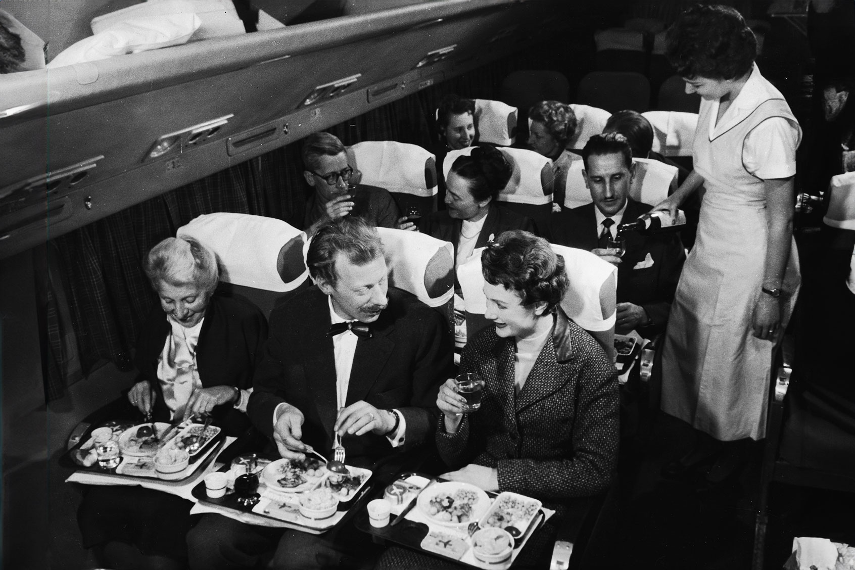 Interior view of a commercial passenger plane shows, in the foreground, a couple as they enjoy their meal next to a smiling elderly woman, while behind them, a flight attendant pours a glass of wine for a man who sits next to a couple who toast each other with full glasses, 1950s. (Frederic Lewis/Getty Images)