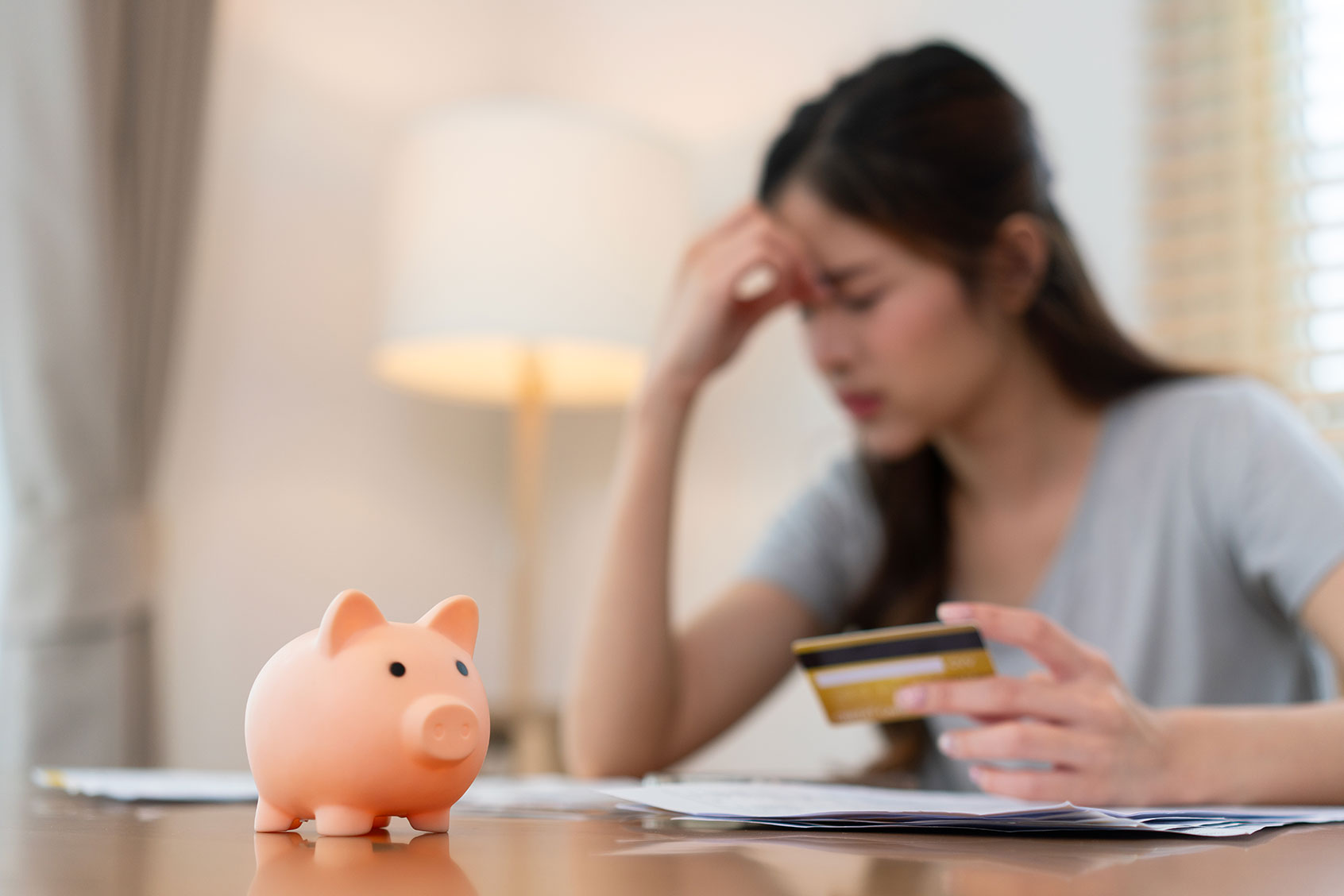 Woman sitting at a desk, holding a credit card and touching her forehead in stress, with a piggy bank in the foreground. (Getty Images/Tfilm)