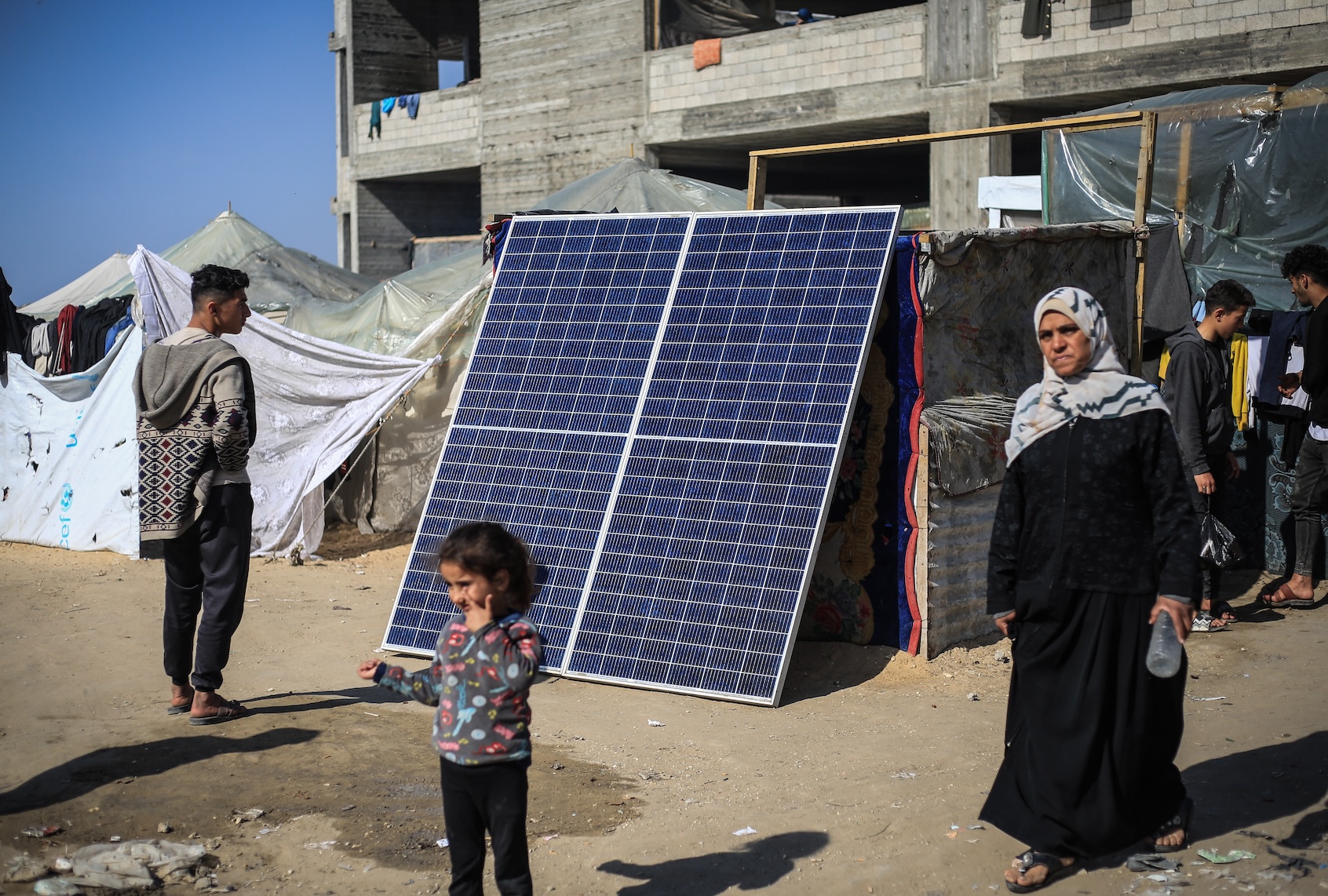 20 January 2024, Palestinian Territories, Rafah: Palestinians walk by a solar panel, used by some to produce electricity in the refugee camps in Rafah. Due to the worsening financial situation of the individuals and the the lack of electricity in the refugee camps in Rafah, some young Palestinian men who have solar panels are using them to run small businesses by which they allow people to fully charge their phones for around 2 Israeli shekels fee (approximately 55 US Cents). (Photo by Mohammed Talatene/picture alliance via Getty Images)