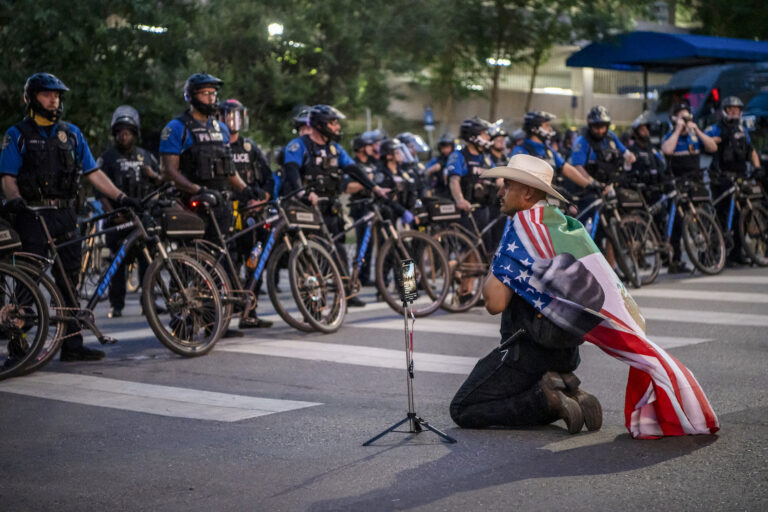 A protester draped with a combined US-Mexican flag kneels in front of a police line while demonstrating against the Trump administration during the "No Kings" national rally in downtown Austin, Texas. (Photo by Sergio Flores/ Getty Images)