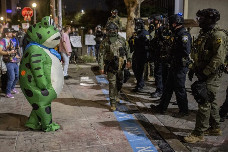 Who are the real Americans? We are Protester in a frog costume before federal officers outside an ICE  facility in Portland, Oregon, Oct. 6, 2025. (Stephen Lam/San Francisco Chronicle via Getty Images)