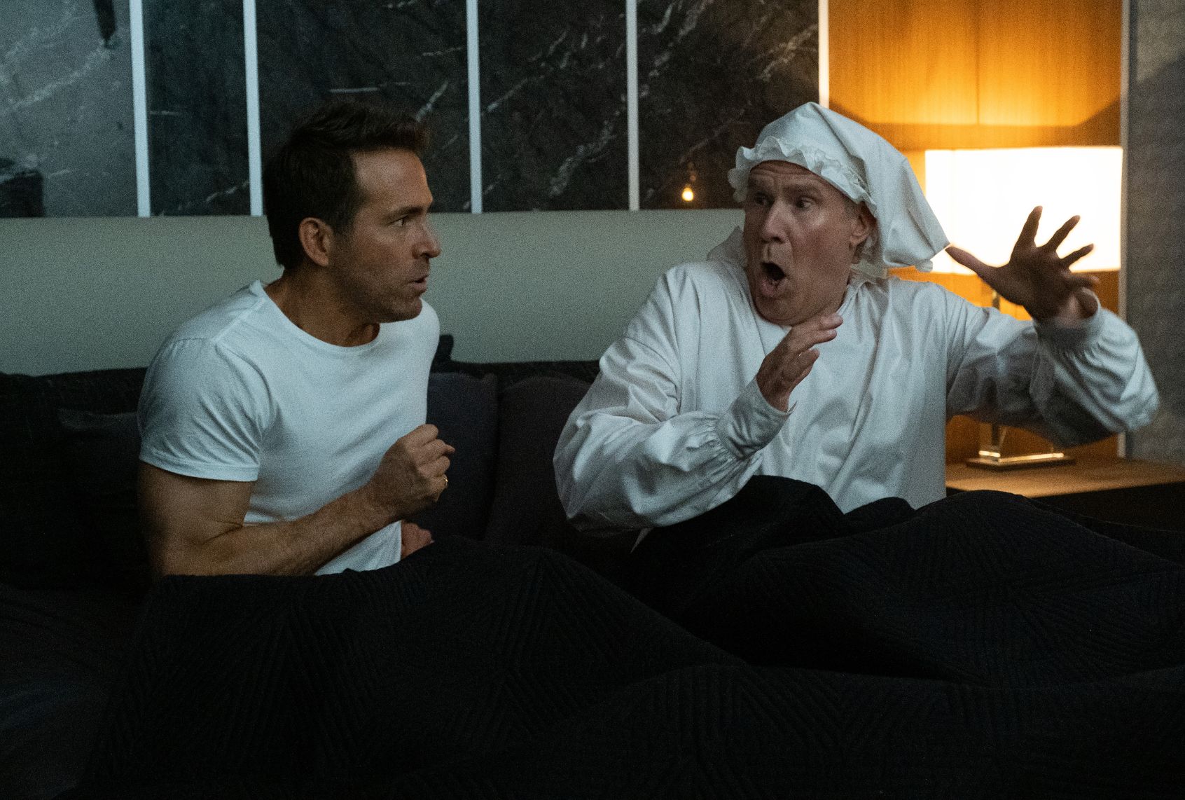 Two men sit upright in bed. One is wearing a white t-shirt and looks concerned, the other is in a white Victorian night shirt with white nightcap on his head and looks startled. A lamp is light on the bedside table