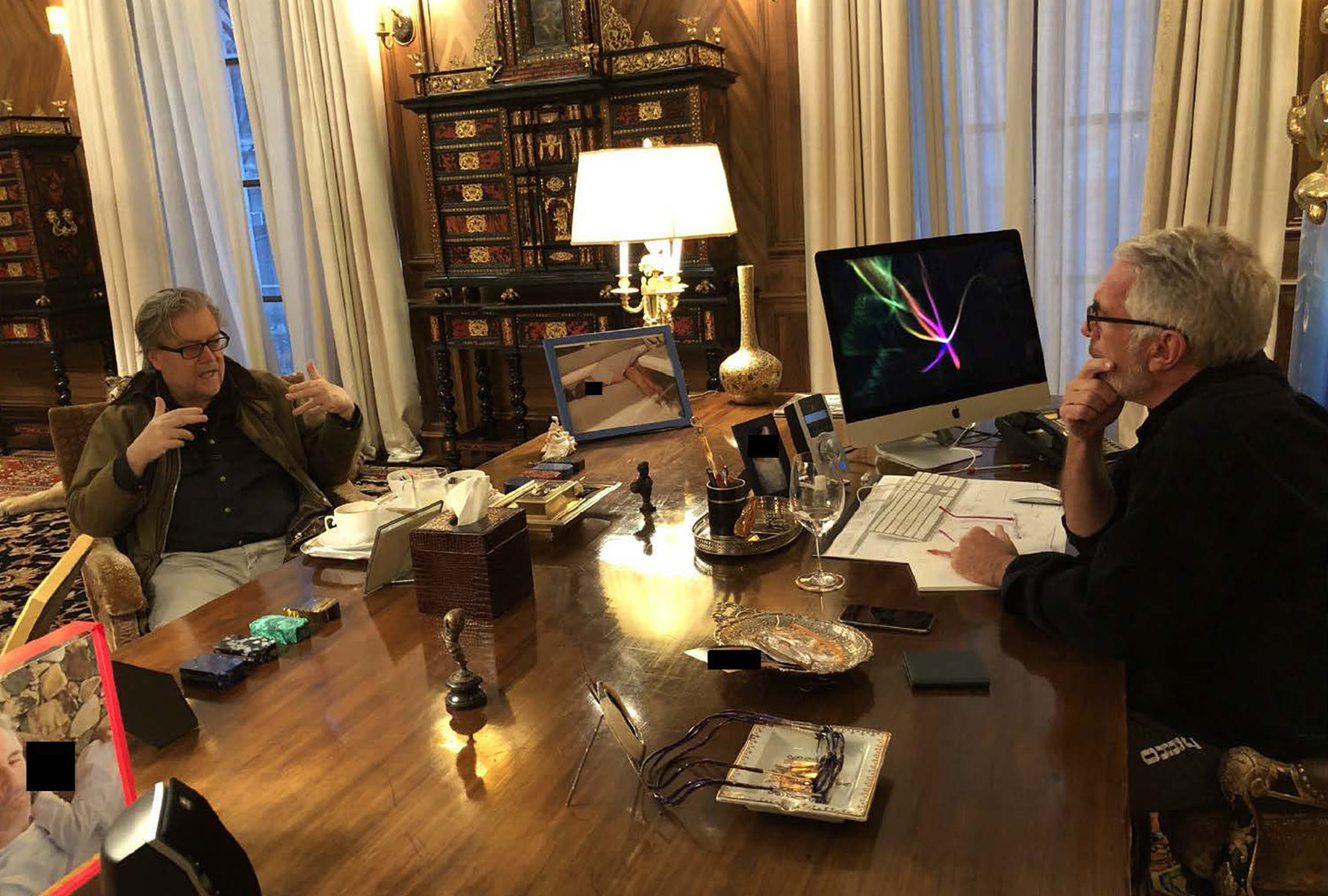 Two men sit across from each other at a large wooden desk during a meeting in an ornate office, with documents, a desktop computer and personal items spread across the table.