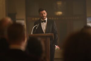 A bearded man in a tux stands in front of a podium to address a crowd
