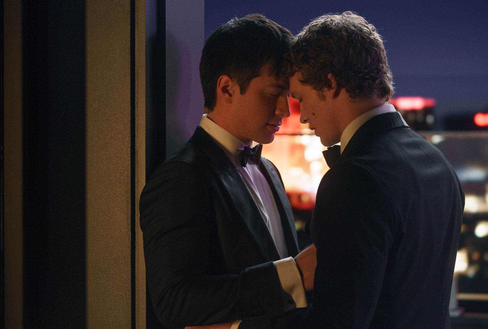 Two men in tuxedos touching foreheads with their eyes closed stand in front of a window