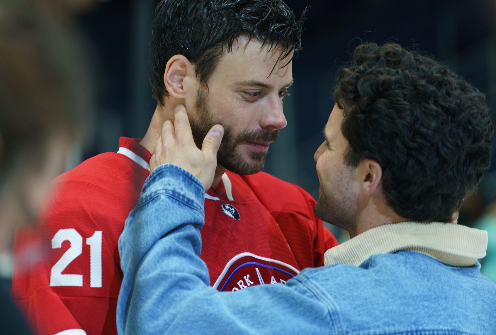 A brunet bearded man in a red hockey jersey gazes gently into the face of a smiling curly-haired man in a denim jacket who is touching the athlete's face gently with one hand
