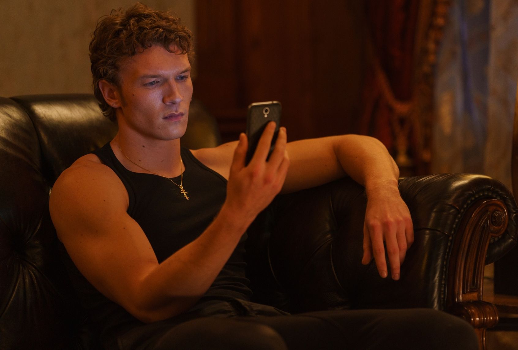 A muscular man wearing black jeans, a black tanktop and silver cross looks at his cell phone with pensive look on his face. He's sitting on an expensive-looking leather couch