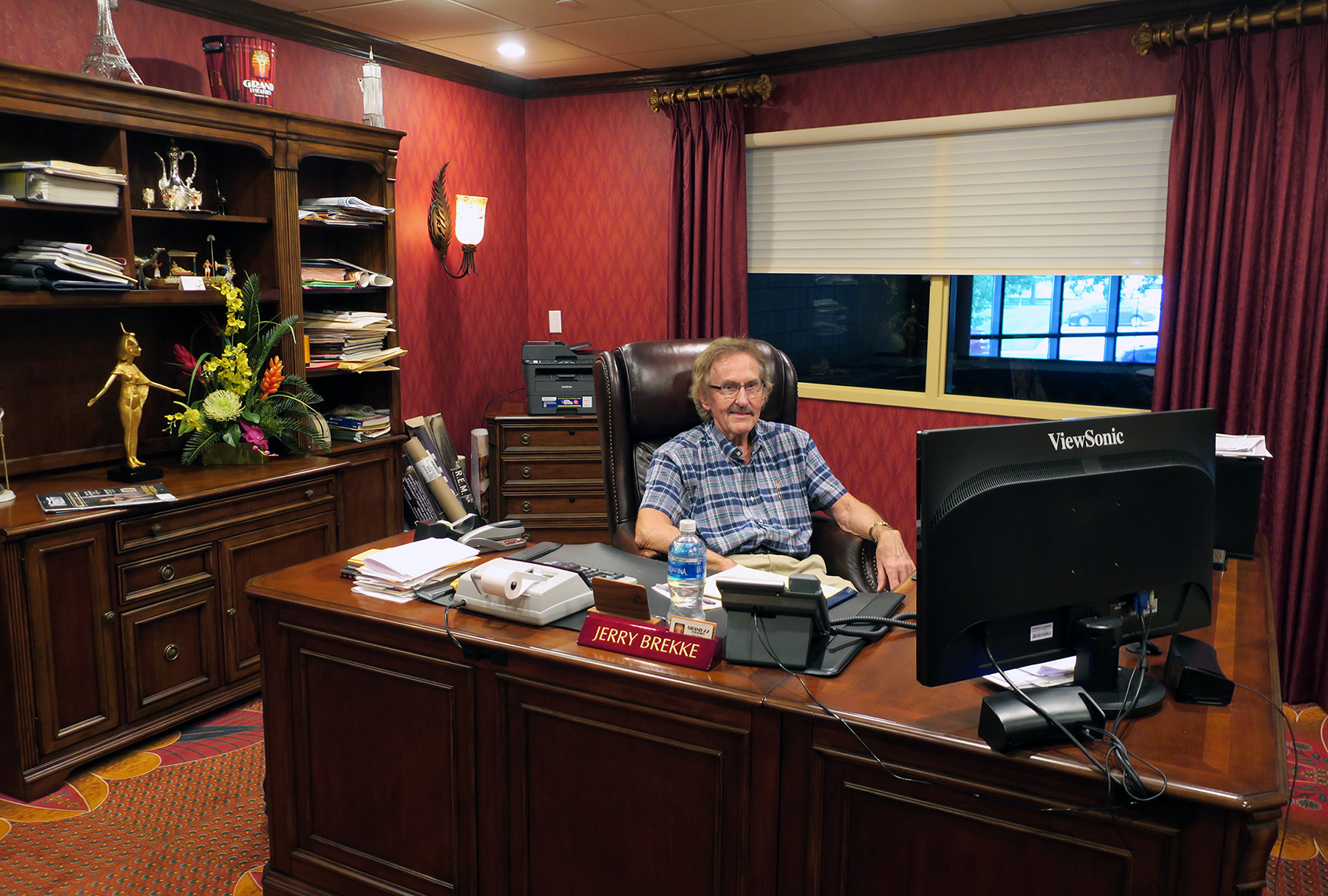 Jerry Brekke sits behind a wooden desk in his office at The Grand in Bismarck, North Dakota, with shelves of papers and decor behind him and a computer monitor in the foreground.
