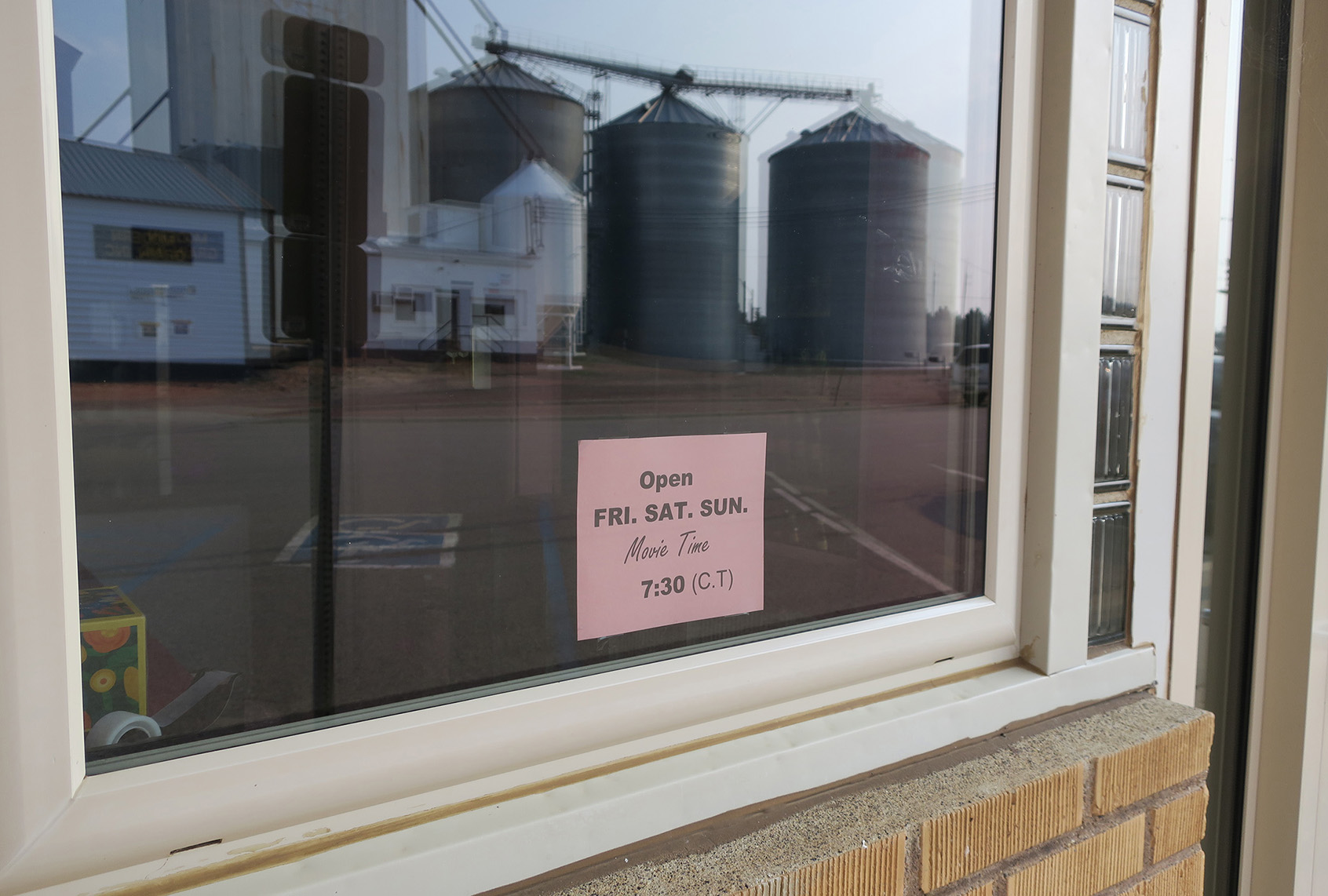 A pink sign taped inside the Mayer Theatre box office window in Hebron, North Dakota, lists movie times as Friday through Sunday at 7:30 p.m., with grain silos reflected in the glass.