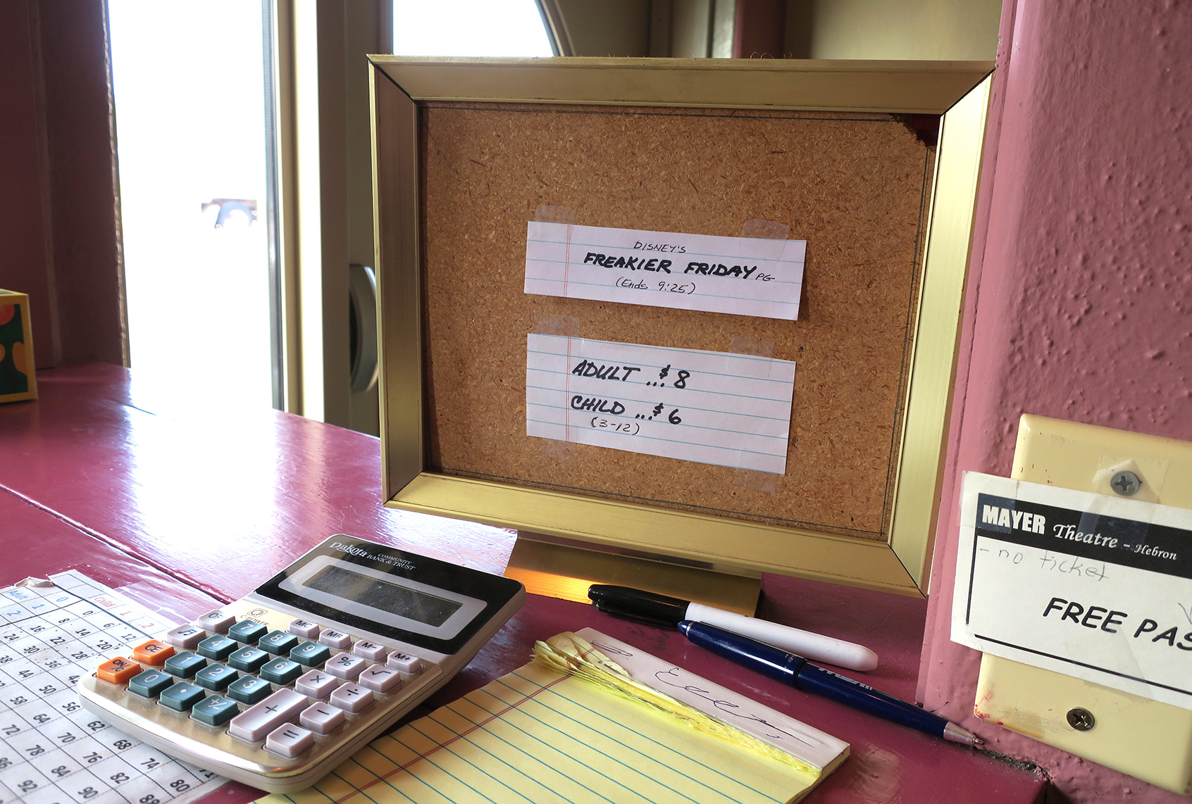 A framed corkboard at the Mayer Theatre box office displays handwritten notes listing a movie title, showtime and adult and child ticket prices, with a calculator and paper on the counter.