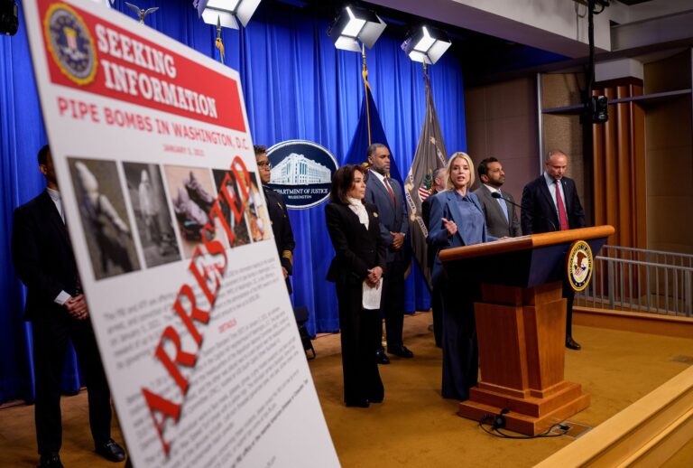 A poster reads "ARRESTED" in bold letters as Attorney General Pam Bondi (3rd-R), accompanied by (L-R) U.S. Attorney for the District of Columbia Jeanine Pirro, Firearms and Explosives (ATF) Washington Field Office Special Agent in Charge Anthony Spotswood, FBI Director Kash Patel, and FBI Deputy Director Dan Bongino, speaks during a news conference on an arrest of a suspect in the January 6th pipe bomb case at the Department of Justice on December 4, 2025 in Washington, DC. (Andrew Harnik/Getty Images)
