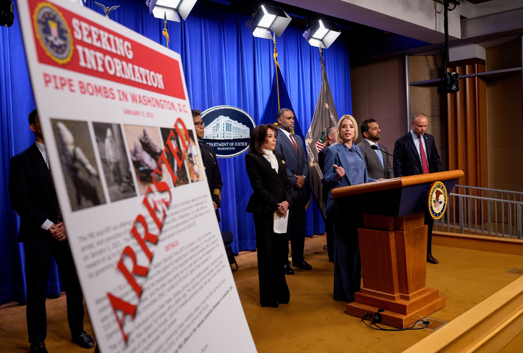 A poster reads "ARRESTED" in bold letters as Attorney General Pam Bondi (3rd-R), accompanied by (L-R) U.S. Attorney for the District of Columbia Jeanine Pirro, Firearms and Explosives (ATF) Washington Field Office Special Agent in Charge Anthony Spotswood, FBI Director Kash Patel, and FBI Deputy Director Dan Bongino, speaks during a news conference on an arrest of a suspect in the January 6th pipe bomb case at the Department of Justice on December 4, 2025 in Washington, DC. (Andrew Harnik/Getty Images)