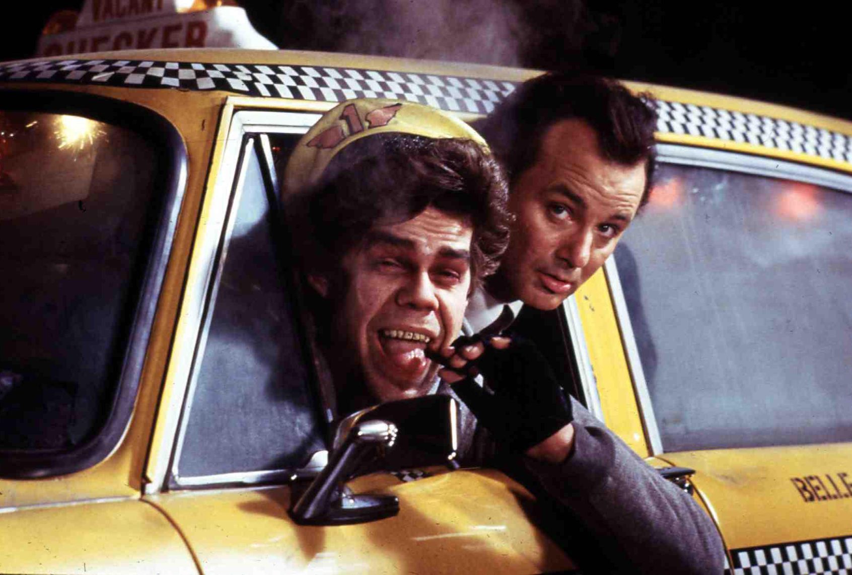 In a classic yellow checker cab, a smiling cabby with a yellow cap and black fingerless gloves sticks his head out of the window as he holds a cigar he has just removed from his mouth. Another brunet man sticks his head out of the same window behind him, also looking out