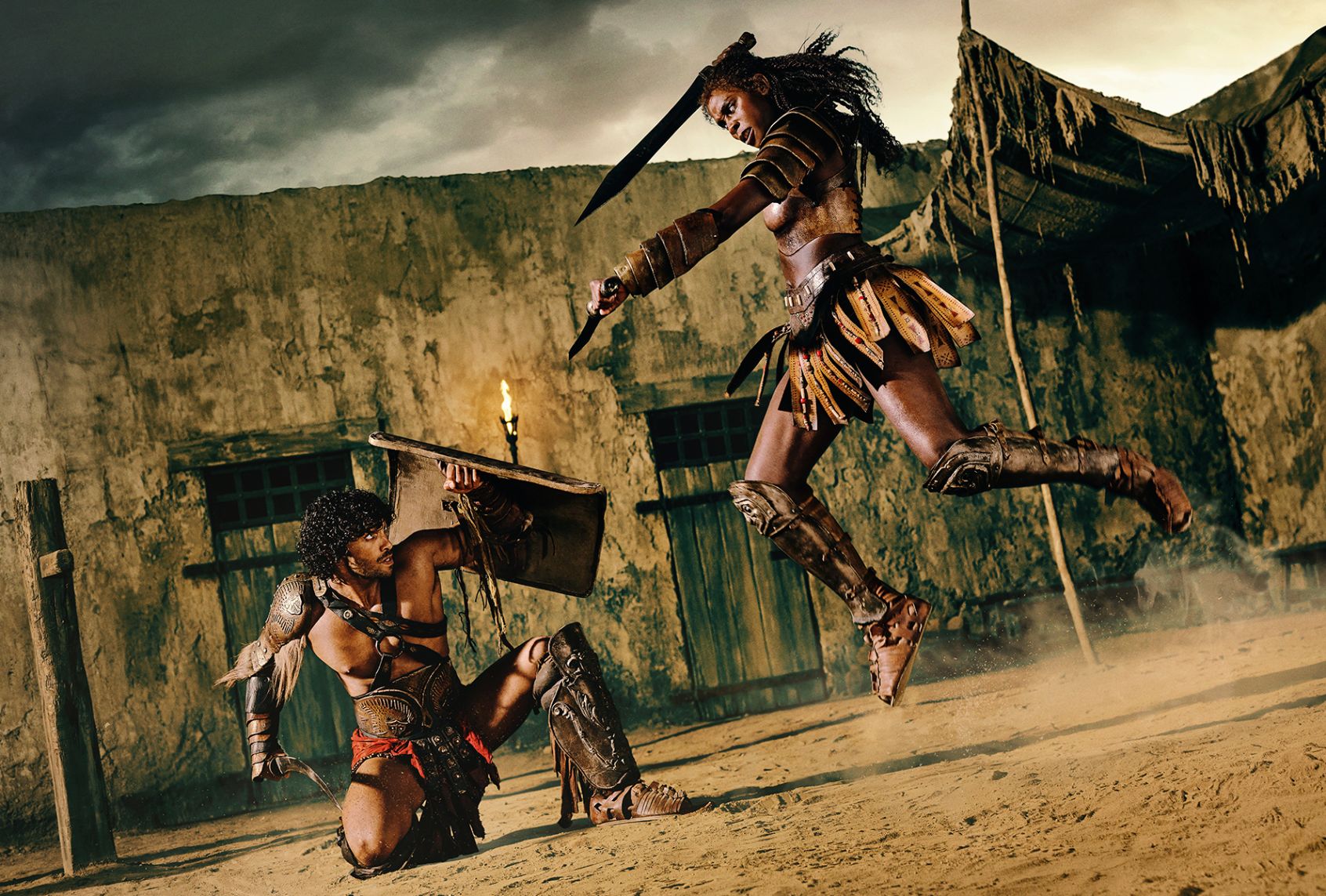 In ancient Rome, a female gladiator in brown leather leaps high, brandishing a sword overhead, about to stab her opponent. The male gladiator defending himself is crouched low on one knee and holds an arched blade in one hand an a shield raised over his head