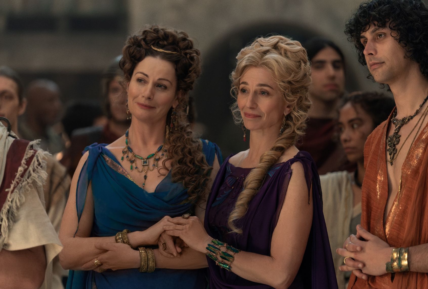 Two women in ancient Rome hold each other's hands in companionship while smiling. One has brown ringlets cascading down her shoulders, a blue tunic and gold diadem with green statement neclace. The other has blond ringlets and is wearing a dark purple gown and green bracelet