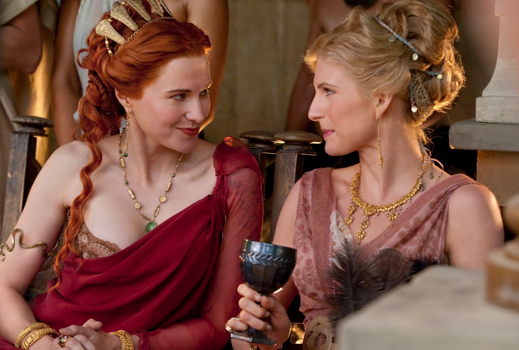 In ancient Rome, two highborn ladies look at each other smling. One has red hair, gold headdress and red draped gown, while the other has her blonde hair piled high, gold jewelry and a dusty pink gown. She is holding a pewter goblet
