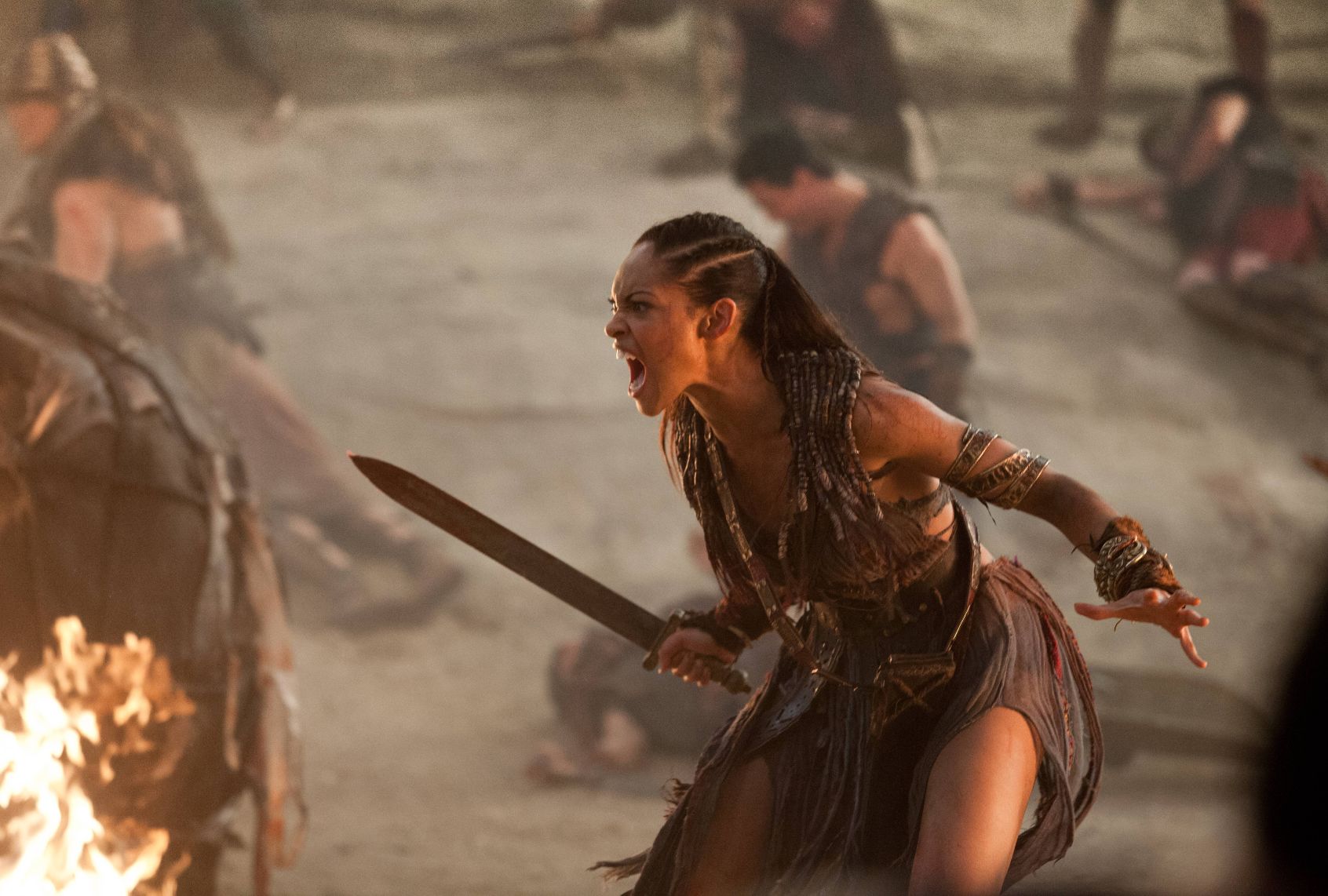 A female gladiator holding a sword is crouched low, screaming and battle ready