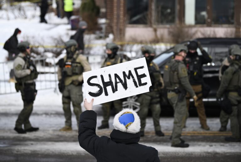 MINNEAPOLIS, MINNESOTA - JANUARY 07: An onlooker holds a sign that reads "Shame" as members of law enforcement work the scene following a suspected shooting by an ICE agent during federal law enforcement operations on January 07, 2026 in Minneapolis, Minnesota. According to federal officials, the agent, “fearing for his life” killed a woman during a confrontation in south Minneapolis. (Photo by Stephen Maturen/Getty Images)