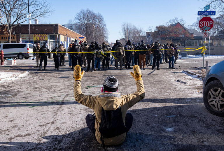 A protester sits on the street with his arms up in front of a gaggle of federal agents and Minneapolis Police on W. 27th St and Nicollet Avenue in south Minneapolis after Alex Pretti was fatally shot by federal agents in the area.