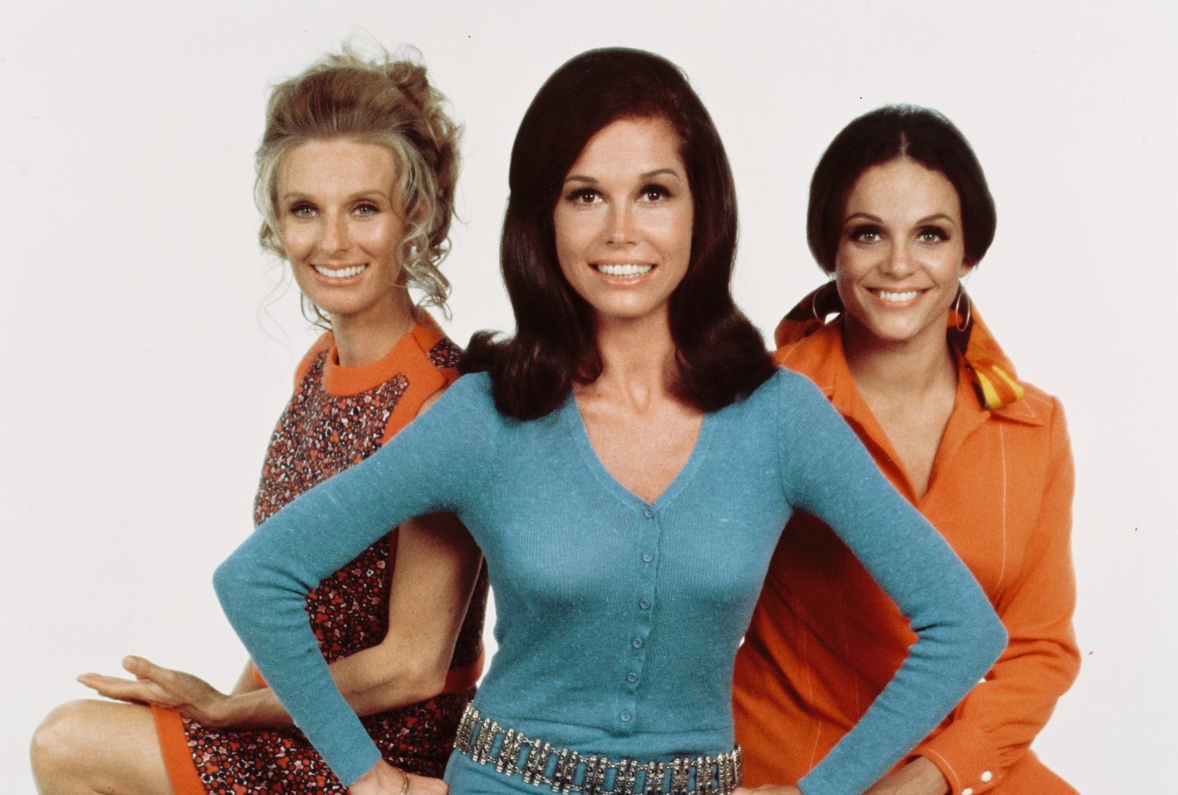 Three women with big smiles face the camera. Two women - one blonde with ringlets and a brunette wearing an orange jumpsuit