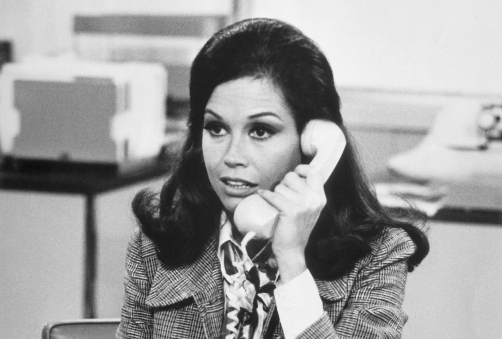 A black & white photo of a 1970s woman with dark bouffant hair and heavy eye makeup. She's wearing a tweed blazer over a print shirt and is talking on the phone, receiver held up to her ear.