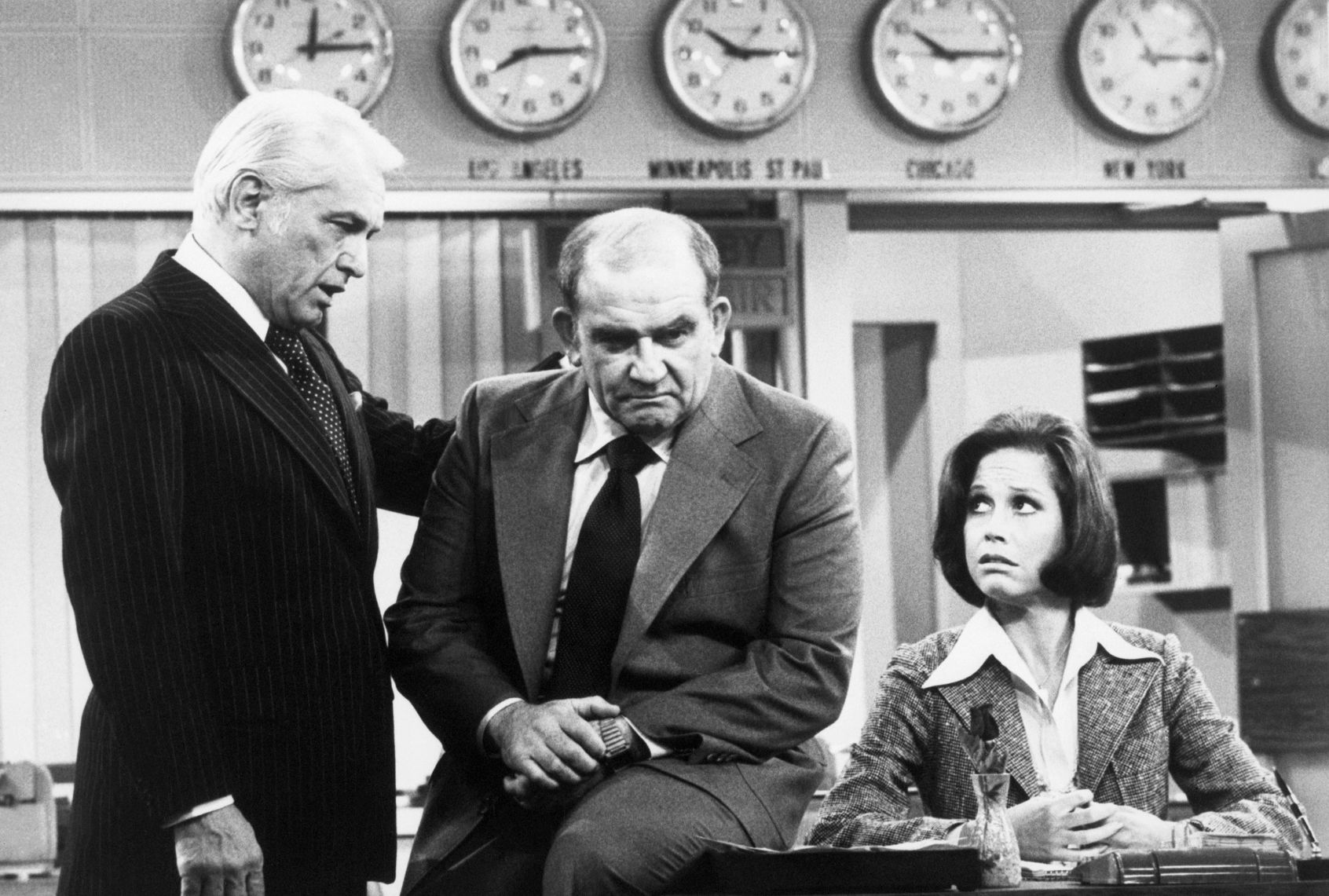 A black & white photo set in a newsroom with a grim balding man in a suit perched on the edge of a desk. The woman - sporting a bob, tweed blazer and big-collared shirt -- is sitting at the desk staring at the man with concern. Another man , a silver-haired blowhard in a dark suit and tie, appears to be consoling the other man with a hand on his shoulder. In the background, six clocks feature various time zones around the country