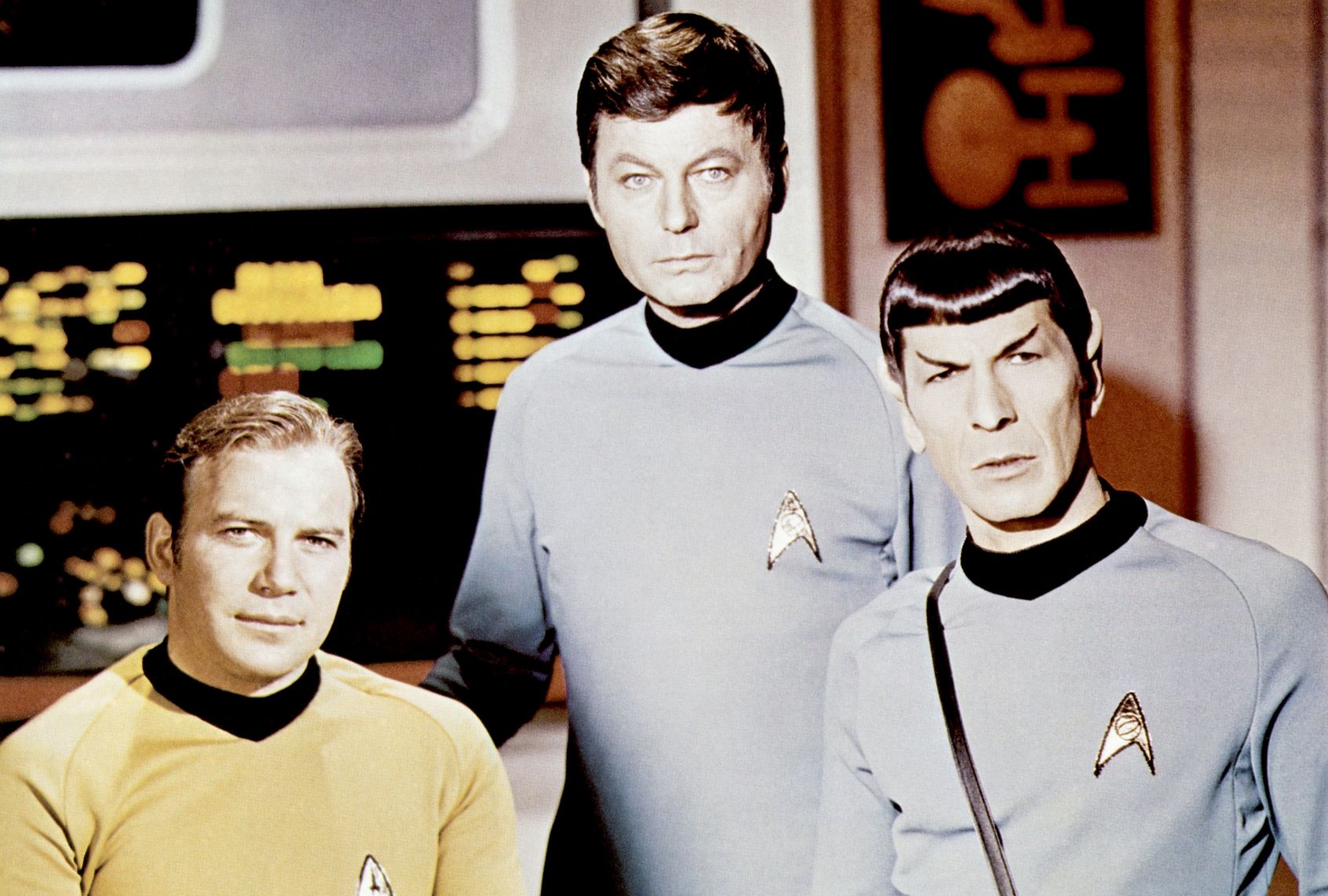 Three stern looking men in form-fitting space uniforms (one in gold, the other in blue) pose in a spaceship filled with computer screens