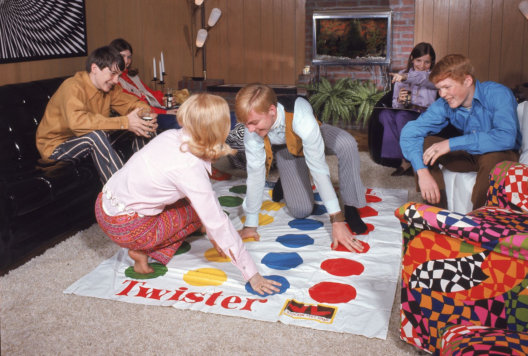 A scene from a 1960s party with teenagers. In a wood-paneled living room, a young man and woman play the party game Twister, with their hands and feet touching colored dots on a white plastic mat. Four other teenagers watch them, laughing. from couches and funky-patterned. chairs.
