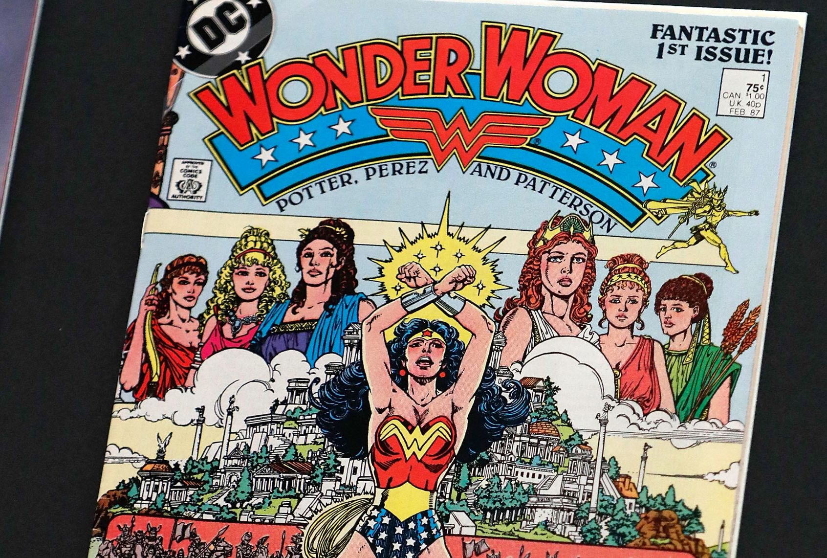A Wonder Woman comic book featuring a woman in red bustier and blue shorts with her arms crossed above her. In the background is a Greek city atop a hill and the image of six Greek women in robes