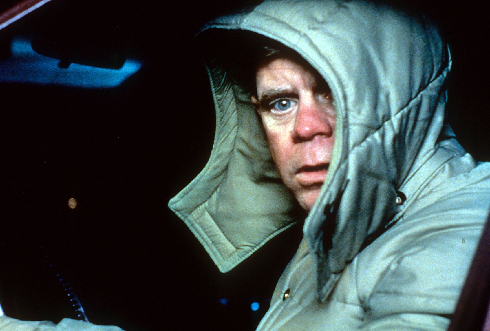 A man with a ruddy complexion and bright blue eyes peeks out from his light green hooded and padded jacket. The interior of a car is dark and shadowed behind him