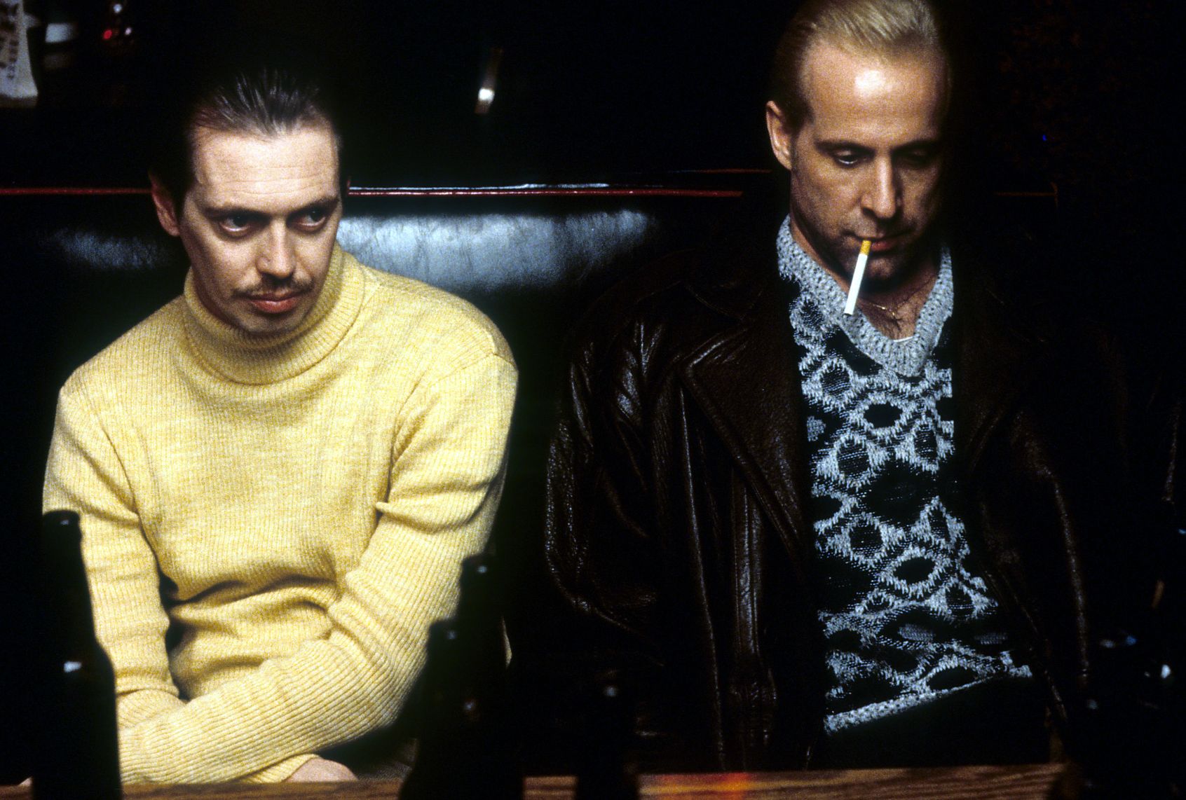 Two men sit in a dark leather booth in a bar. The man on the left has a trim mustache in a sharp face and is wearing a yellow turtleneck sweater. The man on the right wears a dark jacket over a grey and black patterned sweater. An unlit cigarette dangles from his lips