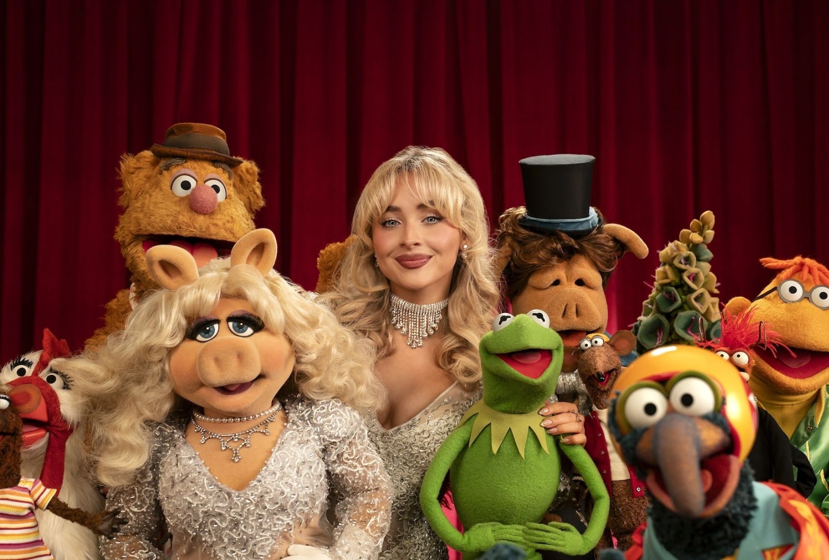 A cast of. felt puppets of different species - bear, pig, frog, chicken, rat and something blue and indeterminate - surround a glamorous blond woman in a pearly white dress and statement choker. Her outfit and bouffant blonde hair is similar to the pig's. They all stand before a red velvet curtain