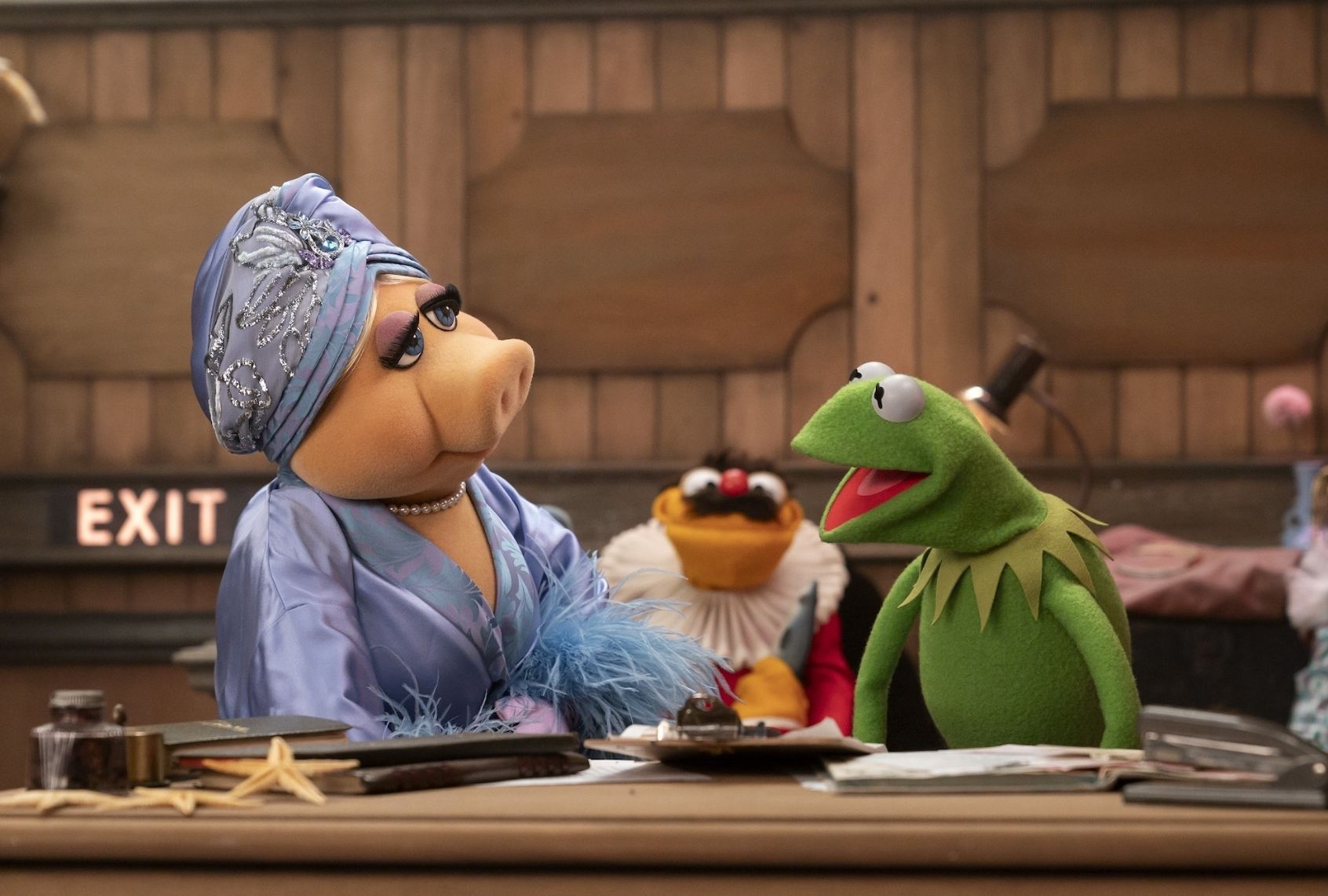 Two puppets - a pretty pig in a lavender gown and headwrap and a frog - have a discussion backstage while an orange puppet with a bushy mustache looks on