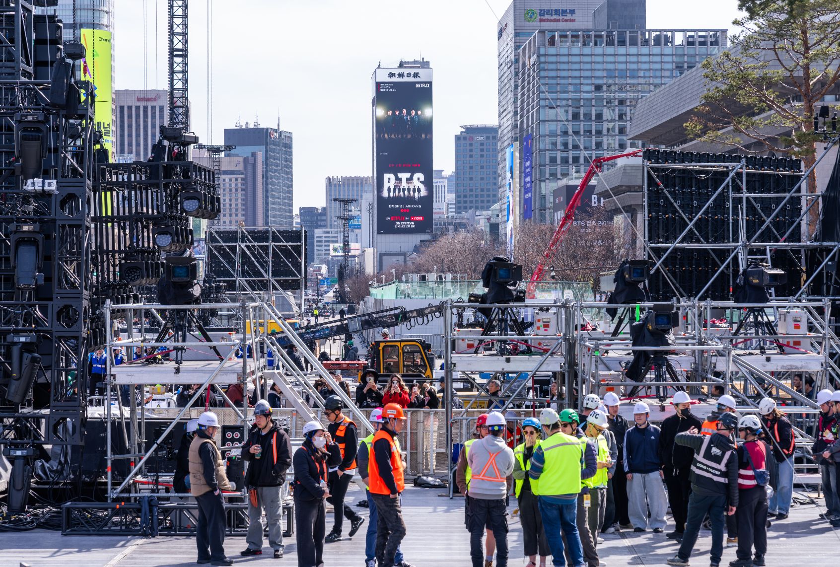 A group of workers in hard hats and safety vests gather around scaffolding and other machines while preparing an outdoor set